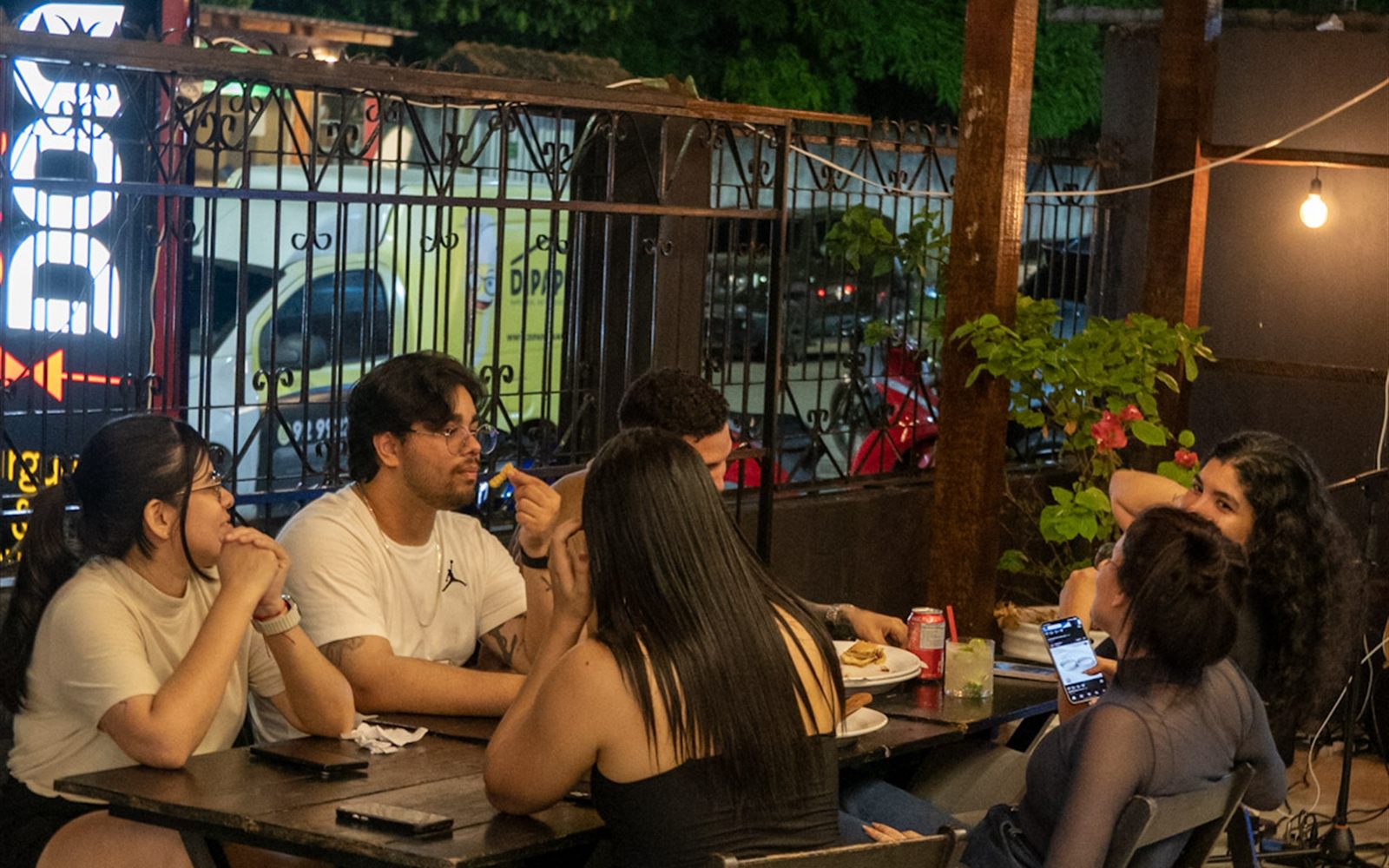 Área externa da casa à noite, com mesas cheias e o letreiro DON na entrada